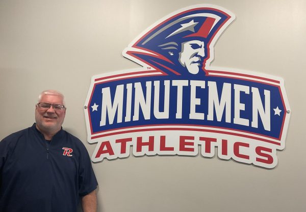 Revere Athletic Director Don Seeker stands beside the Minuteman Athletics sign in his office before retirement.